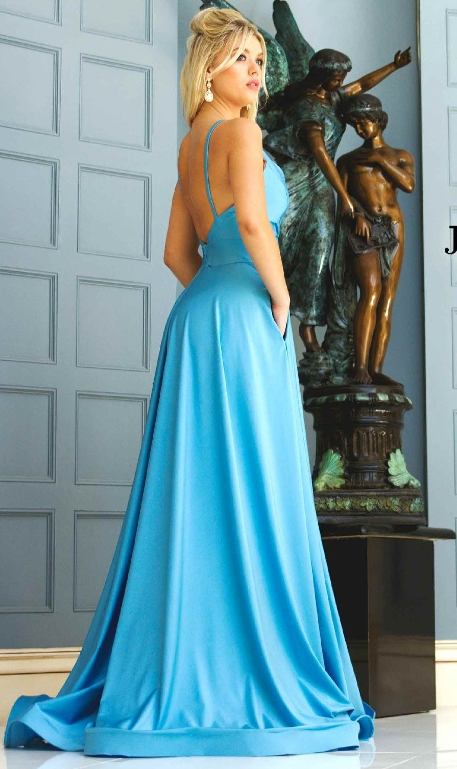 A woman in a light blue, backless Jessica Angel 343 V-Neck Long A-Line Prom Dress stands indoors by a bronze statue and grey paneled wall, looking over her shoulder.